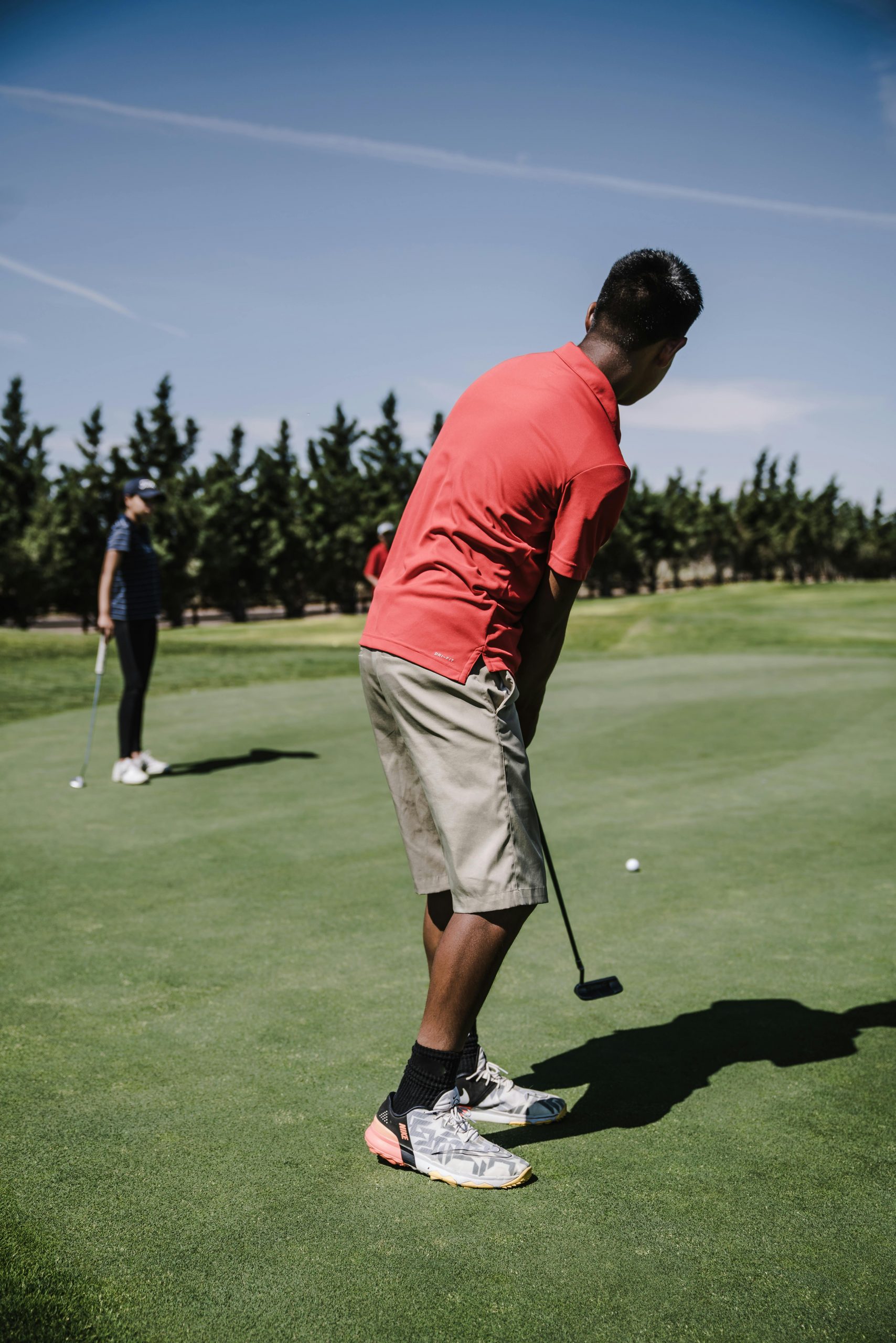 Man in red shirt putting on a green, with another player in background on a sunny day.